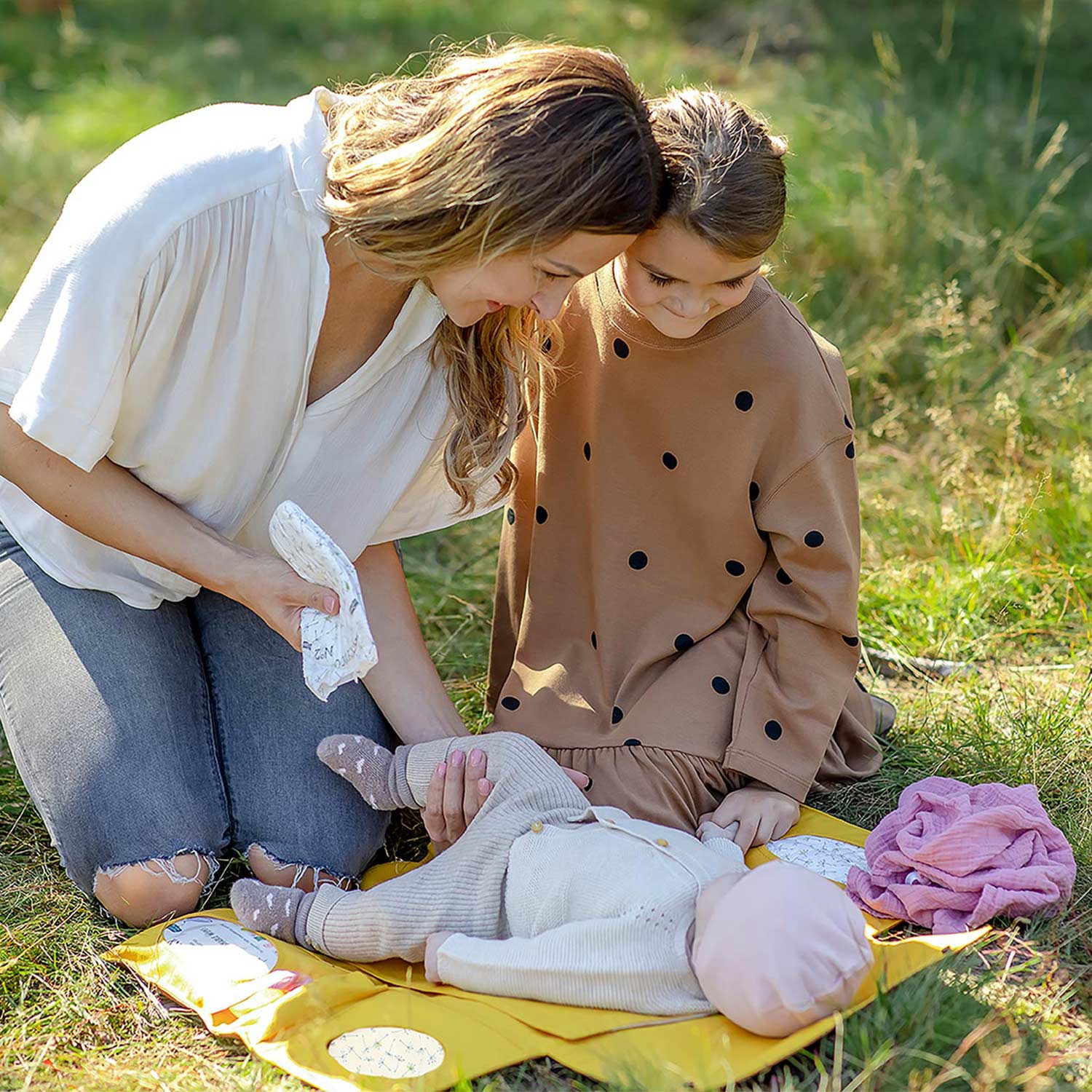 Mutter wickelt Baby zusammen mit älterem Kind auf gelber Villawind Wickelunterlage im Park auf Wiese