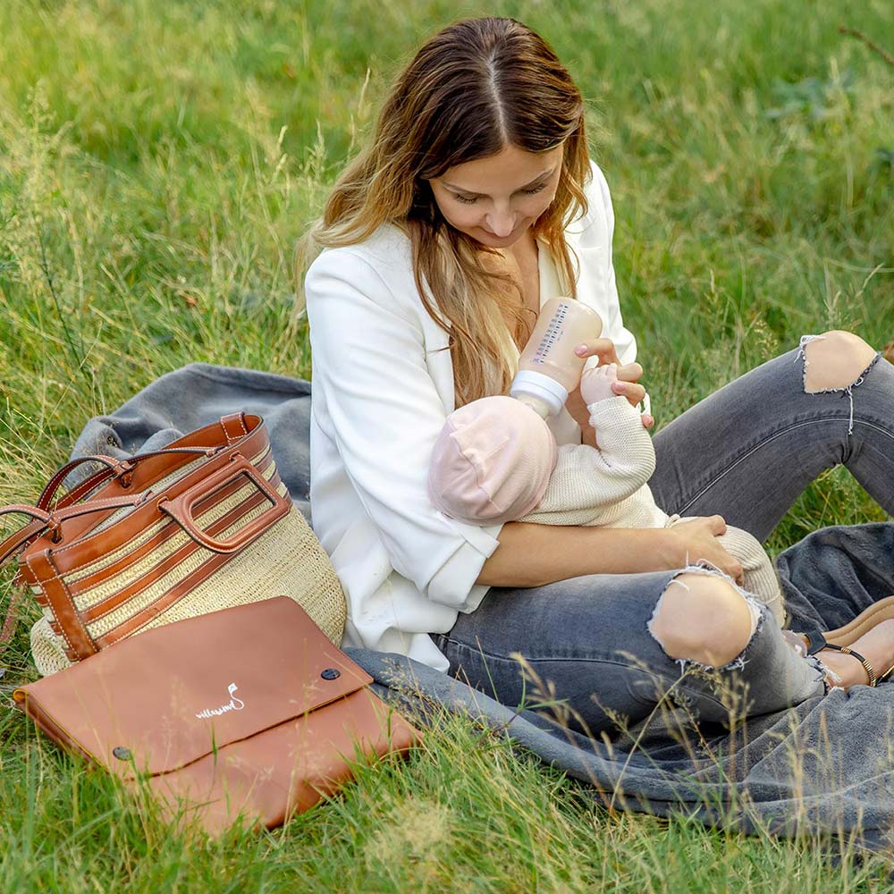 Mutter wickelt Baby auf Villawind Wickeltasche im Park, braune Leder-Wickeltasche neben ihr